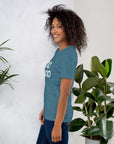 Side view of smiling woman wearing teal Milk Sandwich T-shirt with DO GOOD slogan, standing near indoor plants, minimalist inspirational tee.