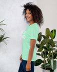 Side view of smiling woman wearing mint Milk Sandwich T-shirt with DO GOOD slogan, standing near indoor plants, minimalist inspirational tee.