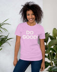 Smiling woman wearing lilac Milk Sandwich T-shirt with distressed white DO GOOD text, standing indoors next to plants, casual inspirational tee.
