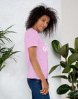 Side profile of smiling woman in lilac Milk Sandwich T-shirt with DO GOOD text, standing near plants, casual inspirational graphic tee.