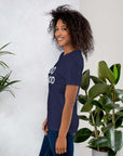 Side view of smiling woman wearing navy Milk Sandwich T-shirt with DO GOOD slogan, standing near indoor plants, minimalist inspirational tee.