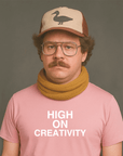 Serious-looking person with glasses, a mustache, a cap with a duck logo, and a mustard scarf, wearing a pink t-shirt that says “HIGH ON CREATIVITY” in white text.