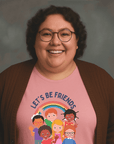 Smiling woman with curly hair and glasses wearing a pink t-shirt that says ‘LET’S BE FRIENDS’ with an illustration of diverse children standing under a rainbow