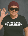 Older woman in sunglasses and beanie wearing “PRETENDING I’M NOT STONED IS MY SUPERPOWER” t-shirt, waving and smiling. Preguntar a ChatGPT