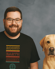 Smiling man with glasses and a beard wearing a black t-shirt that says ‘Round Earth Society’ in colorful text, standing next to a yellow Labrador Retriever dog
