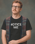 Man wearing glasses and a patterned vest over a black Milk Sandwich t-shirt with bold white distressed text that reads NOTICE ME, looking upward with a curious expression.