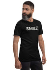 Side-facing man wearing a black Milk Sandwich t-shirt with distressed white text that reads SMILE! on the chest, set against a white backdrop.