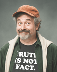 Smiling older man in rust cap and layered outfit wearing black Milk Sandwich T-shirt with bold white text Truth Is Not Fact, humorous statement tee.