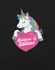 Close-up of a colorful unicorn graphic holding a pink heart with the text “Believe in Edibles” on a black fabric background.