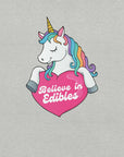 Close-up of a colorful unicorn graphic holding a pink heart with the text “Believe in Edibles” on a light grey fabric background.
