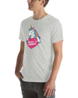 Young man wearing a light grey t-shirt with a colorful unicorn holding a pink heart that says “Believe in Edibles,” standing with hands in pockets, side view.