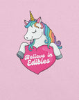 Close-up of a colorful unicorn graphic holding a pink heart with the text “Believe in Edibles” on a lilac fabric background.