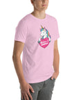 Young man wearing a lilac t-shirt with a colorful unicorn holding a pink heart that says “Believe in Edibles,” standing with hands in pockets, side view.