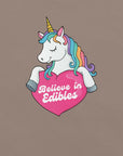 Close-up of a colorful unicorn graphic holding a pink heart with the text “Believe in Edibles” on a pebble fabric background.