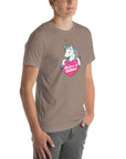 Young man wearing a pebble t-shirt with a colorful unicorn holding a pink heart that says “Believe in Edibles,” standing with hands in pockets, side view.