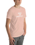 Young man wearing a peach t-shirt with white text ‘HIGH ON CREATIVITY’, standing angled to the side with hands in pockets.