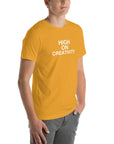 Young man wearing a mustard t-shirt with white text ‘HIGH ON CREATIVITY’, standing angled to the side with hands in pockets.