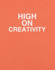 Close-up of orange t-shirt fabric with the phrase “HIGH ON CREATIVITY” printed in bold white uppercase letters in three lines.