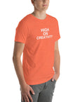 Young man wearing a orange t-shirt with white text ‘HIGH ON CREATIVITY’, standing angled to the side with hands in pockets.