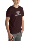 Young man wearing a oxblood black t-shirt with white text ‘HIGH ON CREATIVITY’, standing angled to the side with hands in pockets.