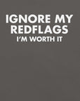 Close-up of asphalt t-shirt with bold white text “IGNORE MY RED FLAGS I’M WORTH IT” printed on the front.
