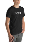 Man wearing black “TOXIC” t-shirt, standing with hands in pockets, looking slightly to the side.