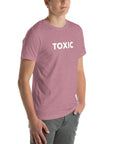 Man wearing orchid “TOXIC” t-shirt, standing with hands in pockets, looking slightly to the side.