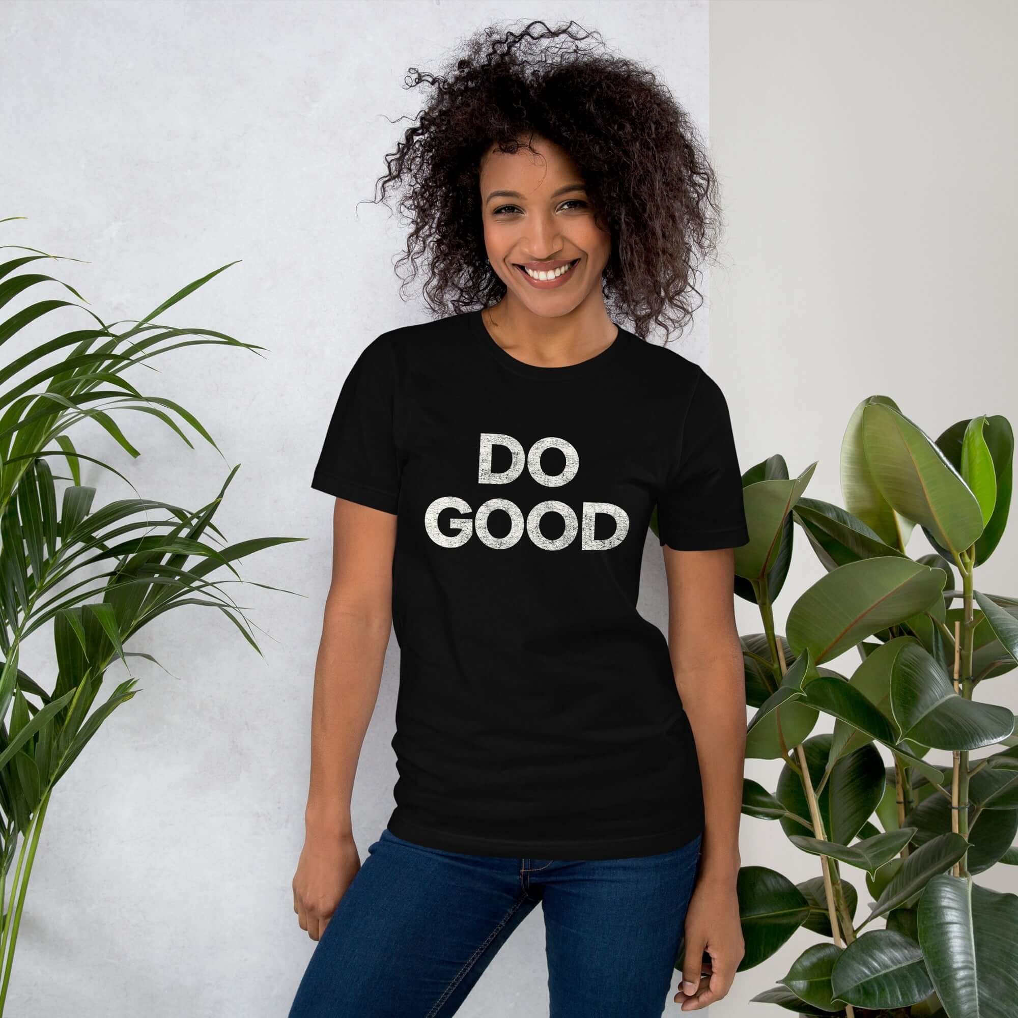 Smiling woman wearing black Milk Sandwich T-shirt with distressed white DO GOOD text, standing indoors next to plants, casual inspirational tee.