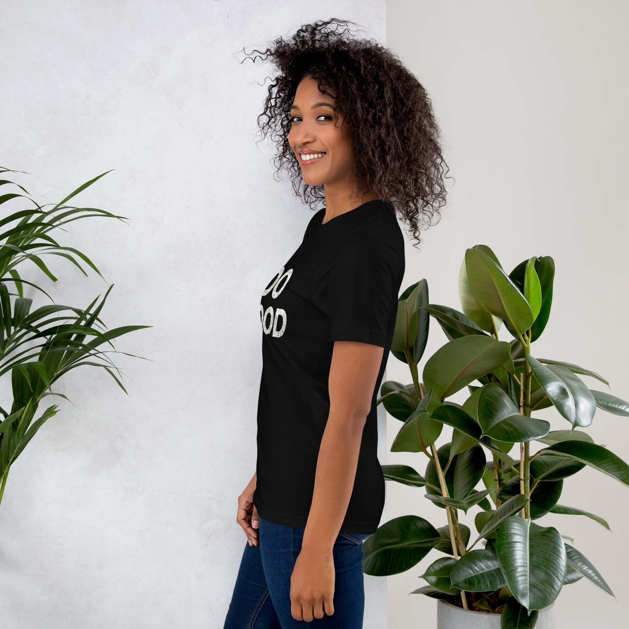 Side view of smiling woman wearing black Milk Sandwich T-shirt with DO GOOD slogan, standing near indoor plants, minimalist inspirational tee.