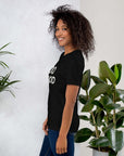 Side view of smiling woman wearing black Milk Sandwich T-shirt with DO GOOD slogan, standing near indoor plants, minimalist inspirational tee.