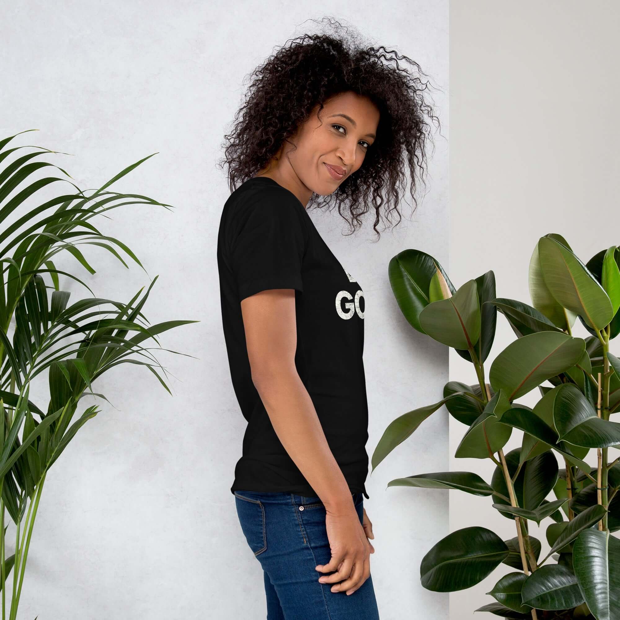 Side profile of smiling woman in black Milk Sandwich T-shirt with DO GOOD text, standing near plants, casual inspirational graphic tee.