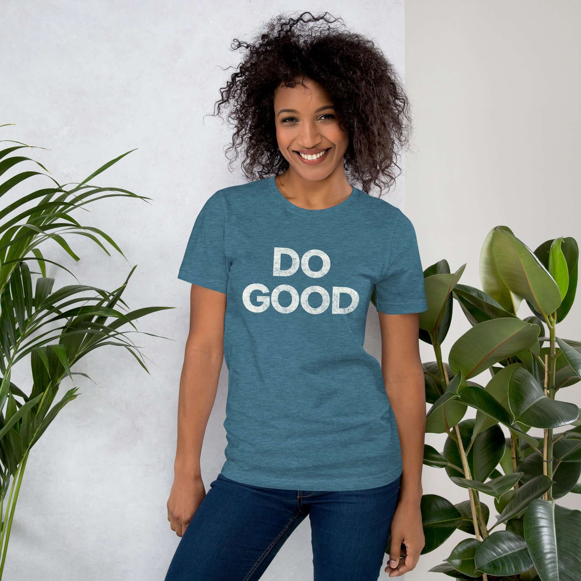 Smiling woman wearing teal Milk Sandwich T-shirt with distressed white DO GOOD text, standing indoors next to plants, casual inspirational tee.