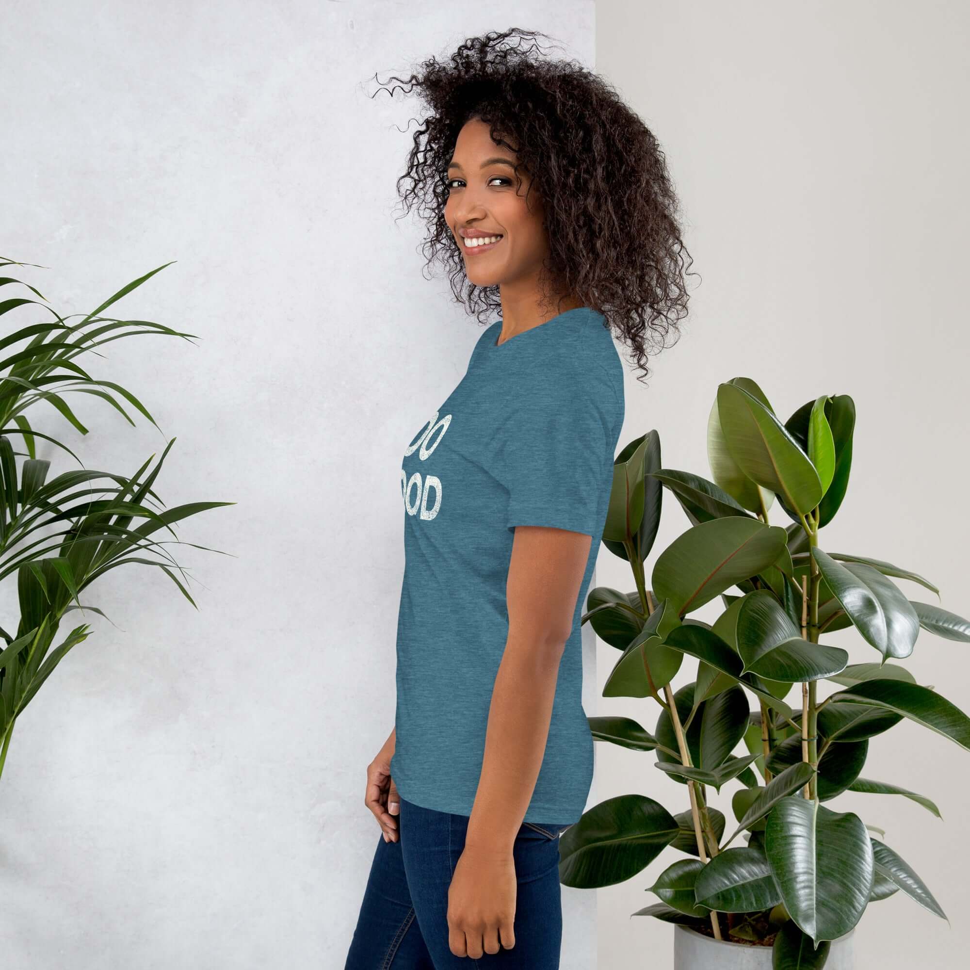 Side view of smiling woman wearing teal Milk Sandwich T-shirt with DO GOOD slogan, standing near indoor plants, minimalist inspirational tee.