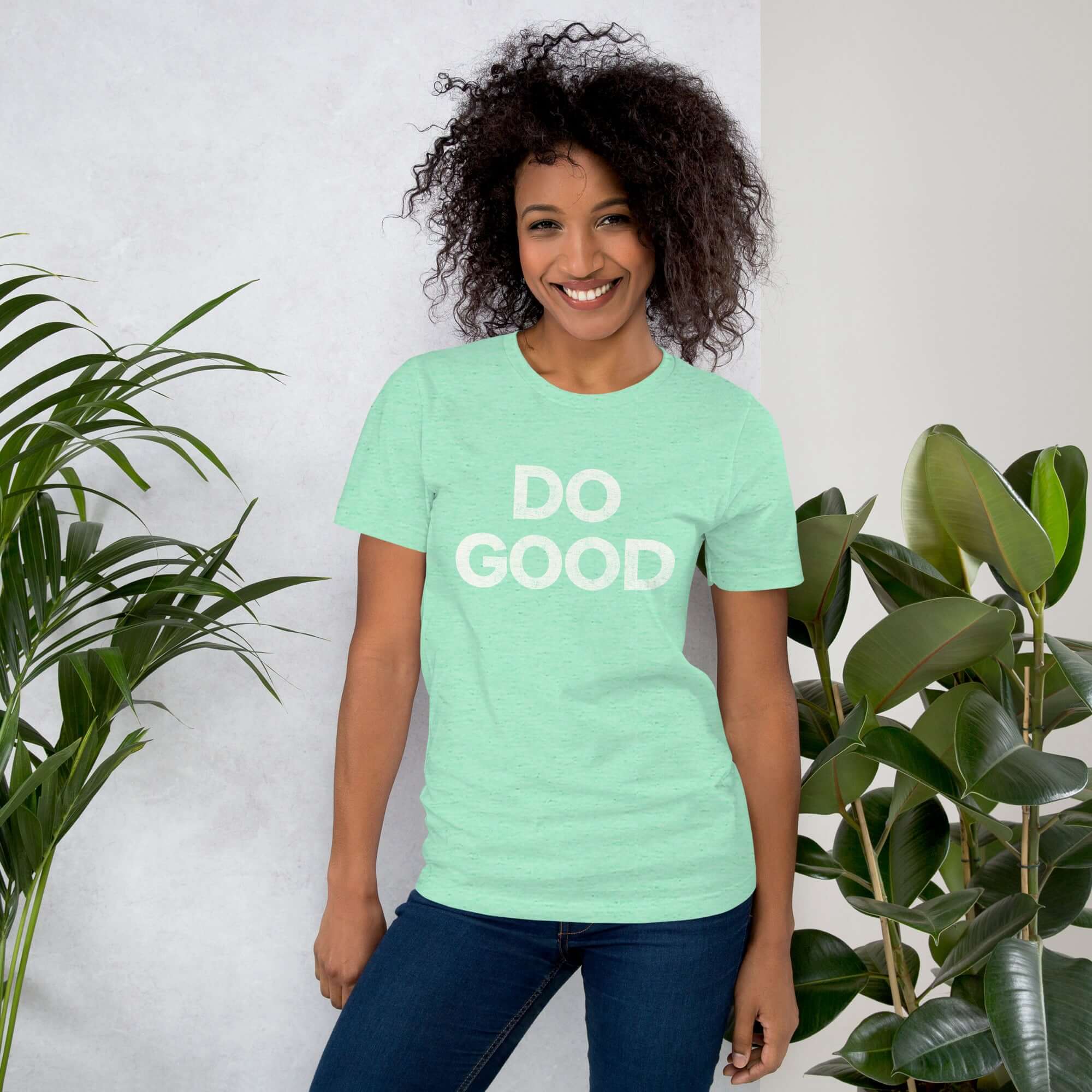 Smiling woman wearing heather mint Milk Sandwich T-shirt with distressed white DO GOOD text, standing indoors next to plants, casual inspirational tee.