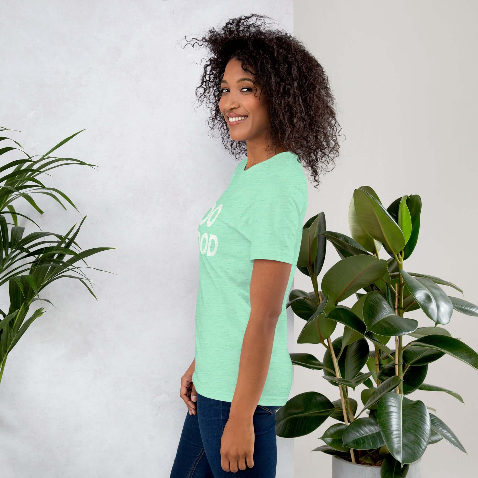 Side view of smiling woman wearing mint Milk Sandwich T-shirt with DO GOOD slogan, standing near indoor plants, minimalist inspirational tee.