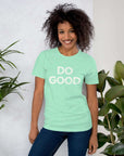 Smiling woman wearing heather mint Milk Sandwich T-shirt with distressed white DO GOOD text, standing indoors next to plants, casual inspirational tee.