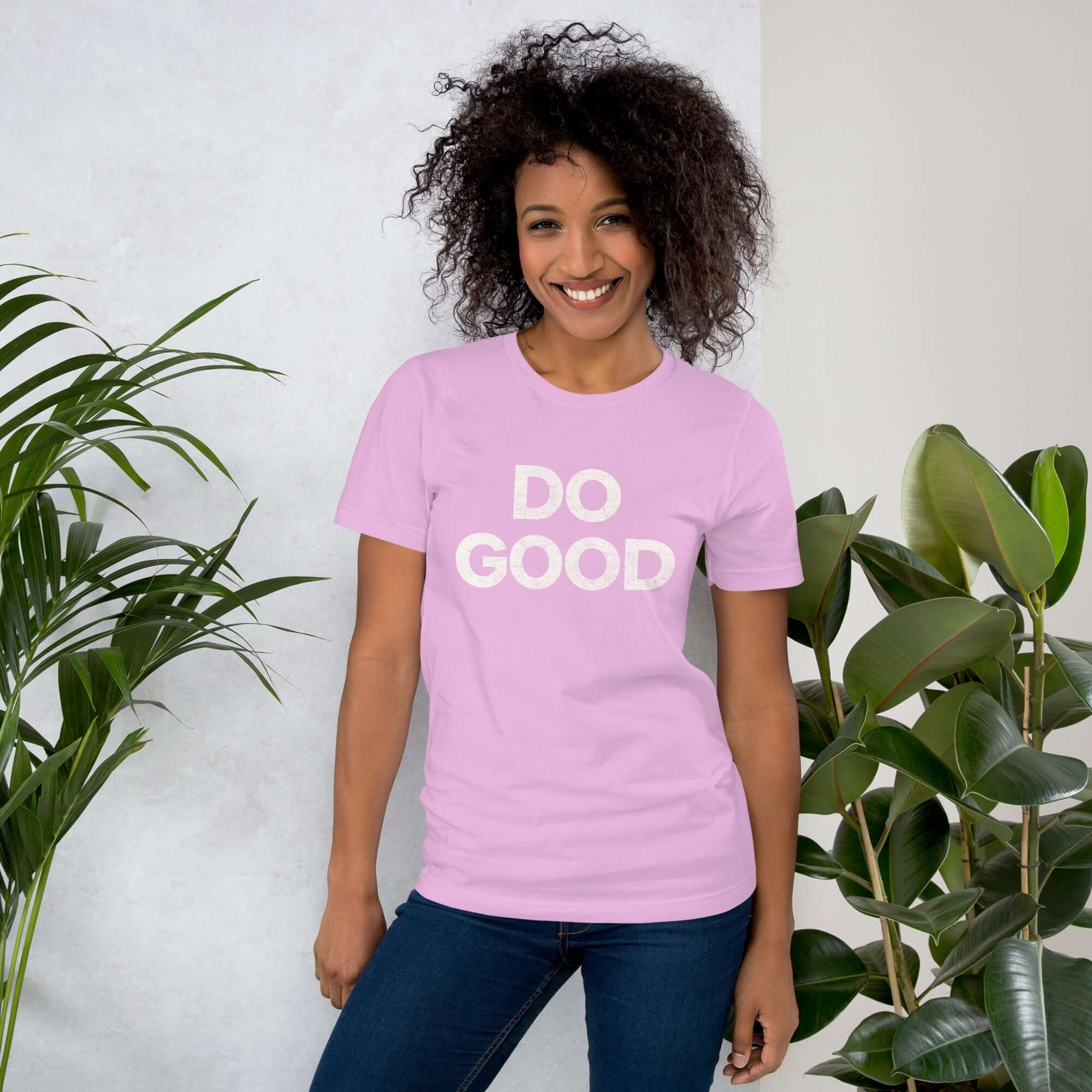 Smiling woman wearing lilac Milk Sandwich T-shirt with distressed white DO GOOD text, standing indoors next to plants, casual inspirational tee.