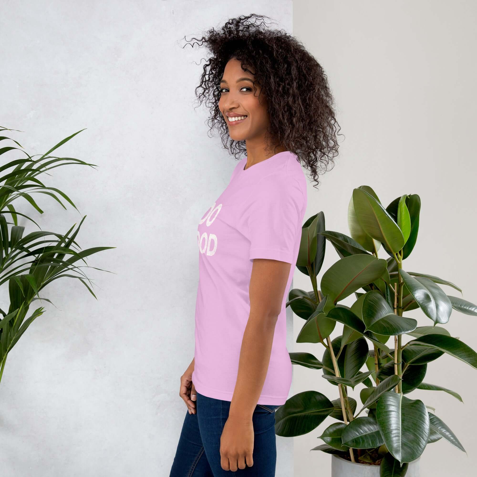 Side view of smiling woman wearing lilac Milk Sandwich T-shirt with DO GOOD slogan, standing near indoor plants, minimalist inspirational tee.