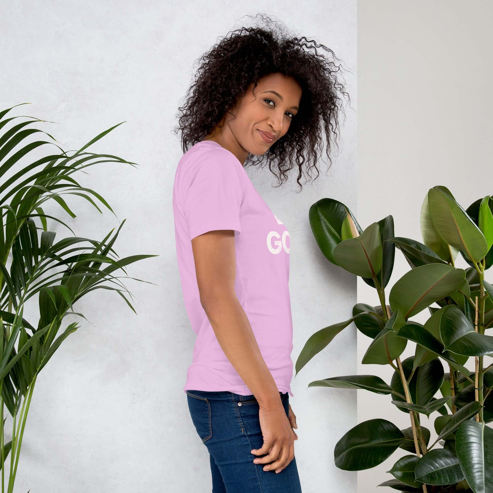 Side profile of smiling woman in lilac Milk Sandwich T-shirt with DO GOOD text, standing near plants, casual inspirational graphic tee.
