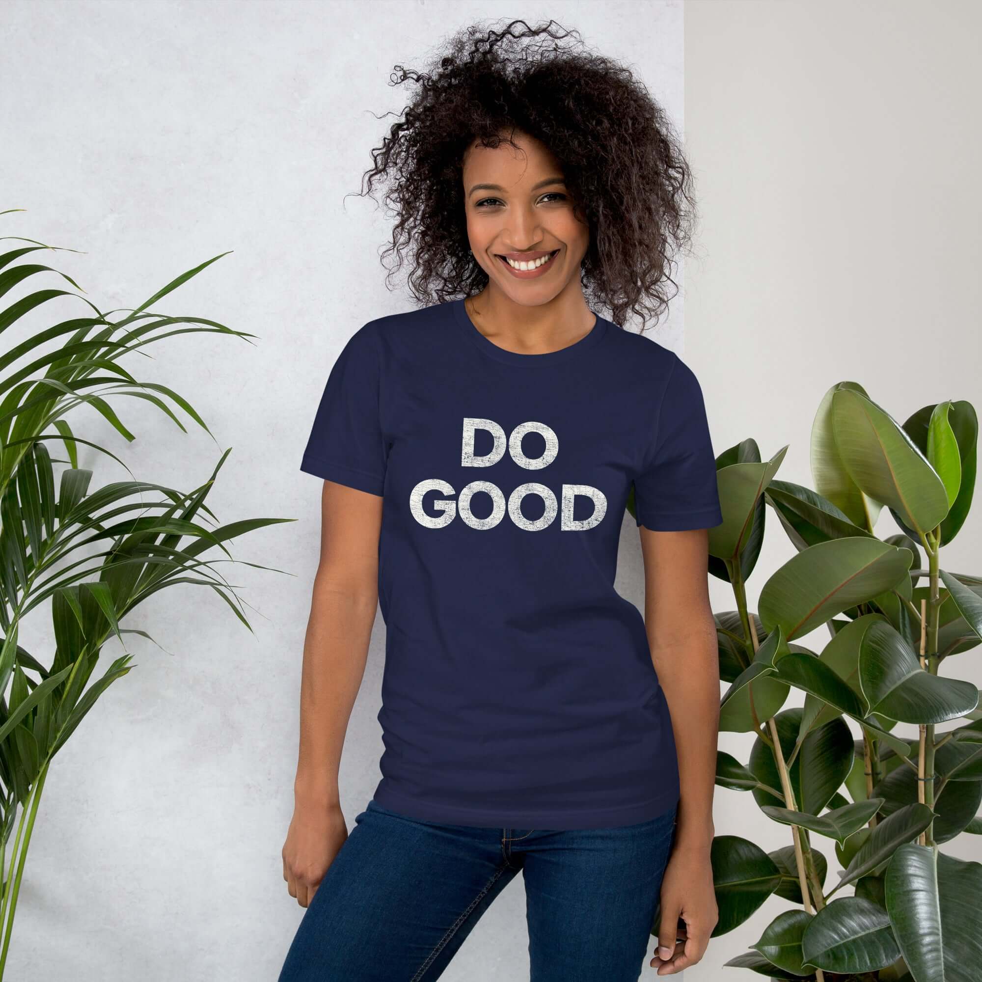 Smiling woman wearing navy Milk Sandwich T-shirt with distressed white DO GOOD text, standing indoors next to plants, casual inspirational tee.