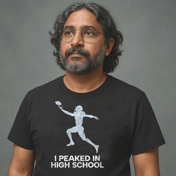 Man with glasses and long hair wearing black Milk Sandwich T-shirt with distressed football player graphic and text I Peaked in High School, nostalgic athletic tee.