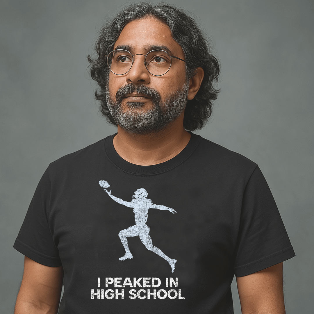 Man with glasses and long hair wearing black Milk Sandwich T-shirt with distressed football player graphic and text I Peaked in High School, nostalgic athletic tee.