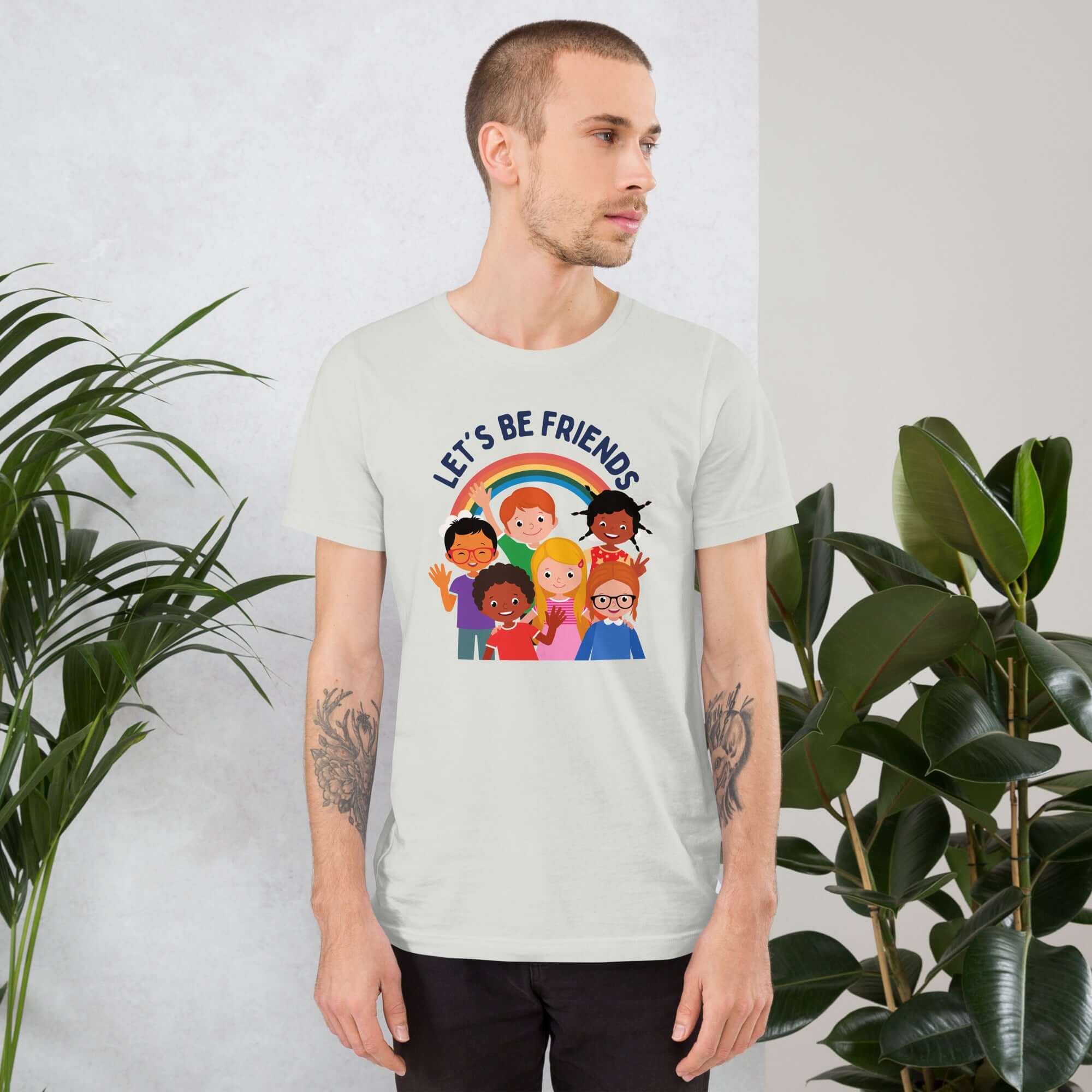 Man wearing a ash Milk Sandwich t-shirt featuring a colorful illustration of six diverse cartoon kids smiling under a rainbow with the text "LET'S BE FRIENDS" standing indoors next to large green plants.