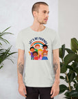 Man wearing a ash Milk Sandwich t-shirt featuring a colorful illustration of six diverse cartoon kids smiling under a rainbow with the text "LET'S BE FRIENDS" standing indoors next to large green plants.