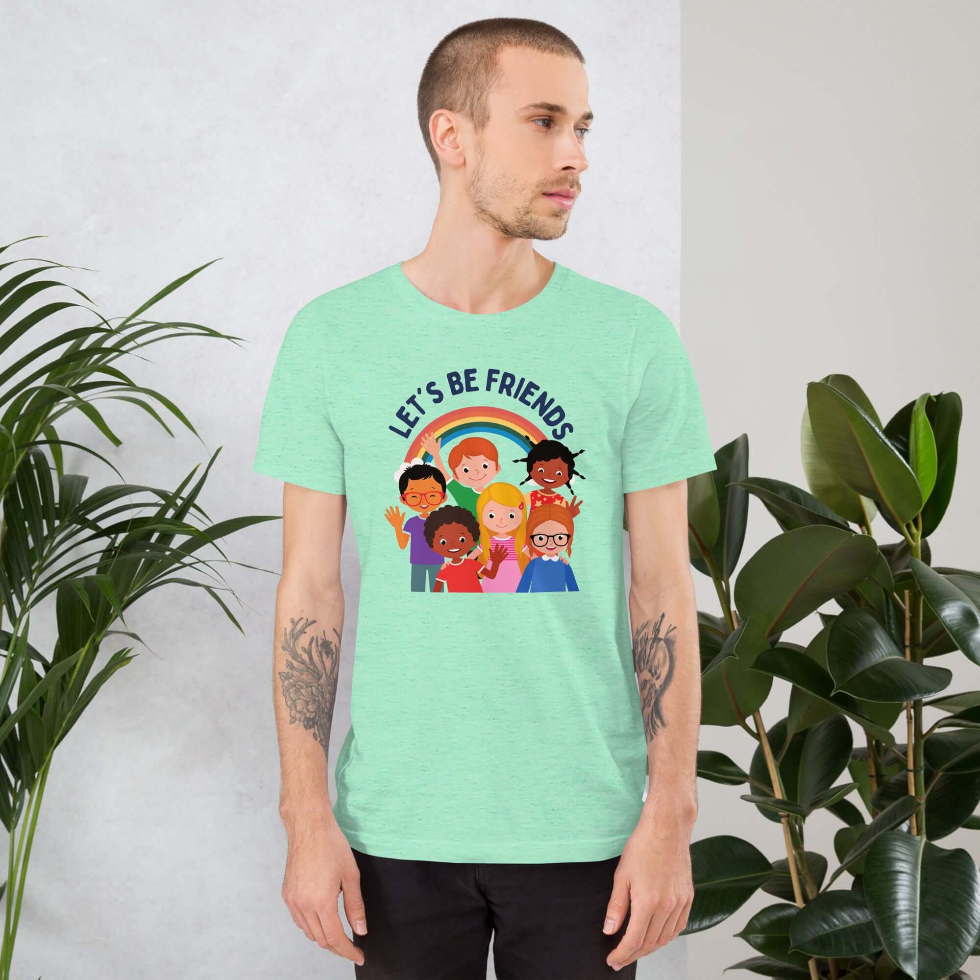 Man wearing a light mint Milk Sandwich t-shirt featuring a colorful illustration of six diverse cartoon kids smiling under a rainbow with the text "LET'S BE FRIENDS" standing indoors next to large green plants.