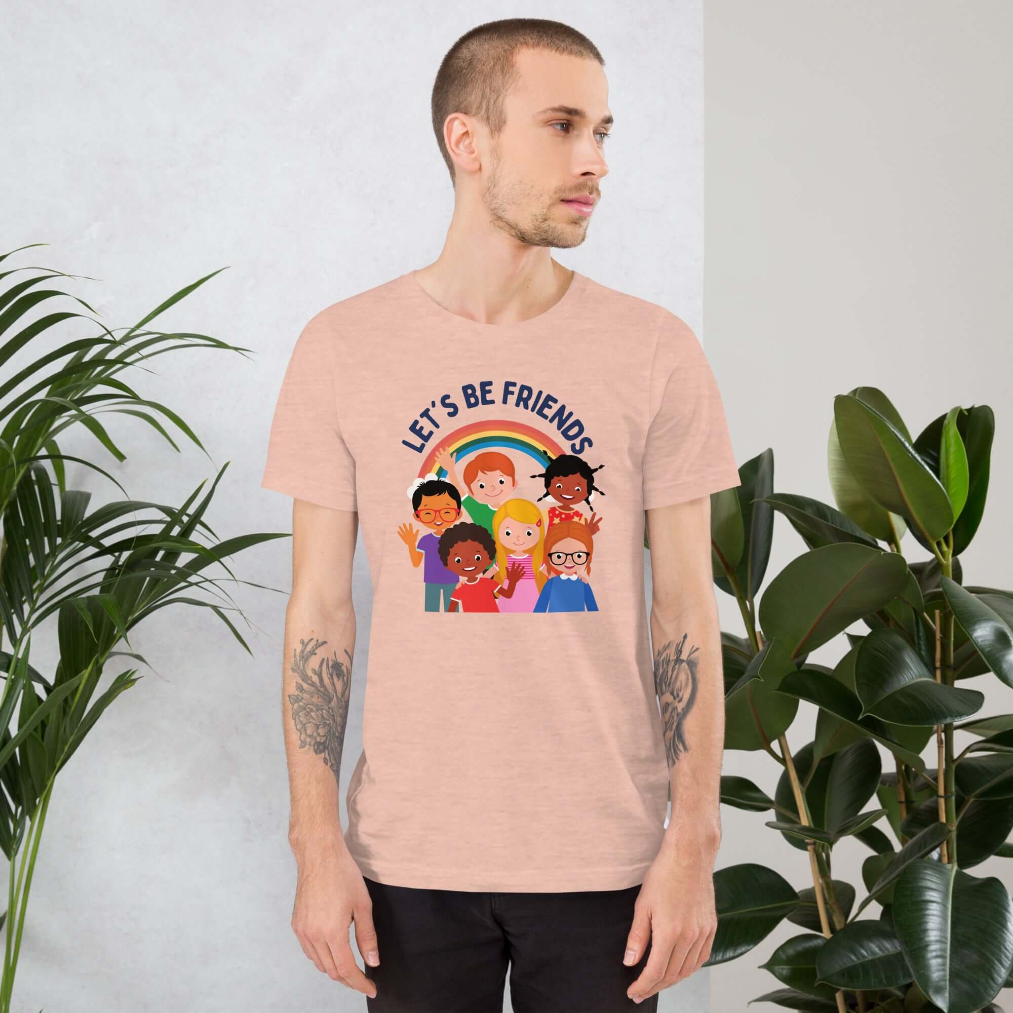 Man wearing a light peachy Milk Sandwich t-shirt featuring a colorful illustration of six diverse cartoon kids smiling under a rainbow with the text "LET'S BE FRIENDS" standing indoors next to large green plants.