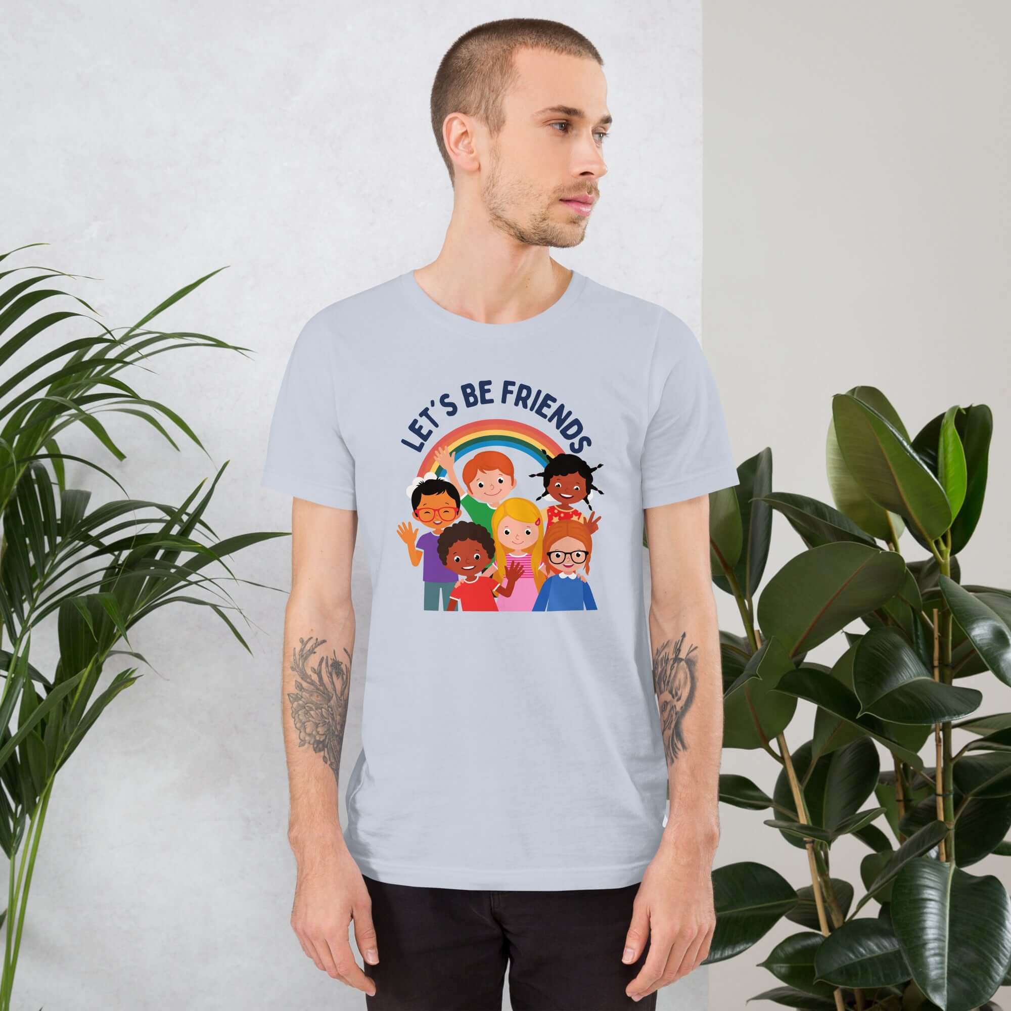 Man wearing a light blue Milk Sandwich t-shirt featuring a colorful illustration of six diverse cartoon kids smiling under a rainbow with the text "LET'S BE FRIENDS" standing indoors next to large green plants.