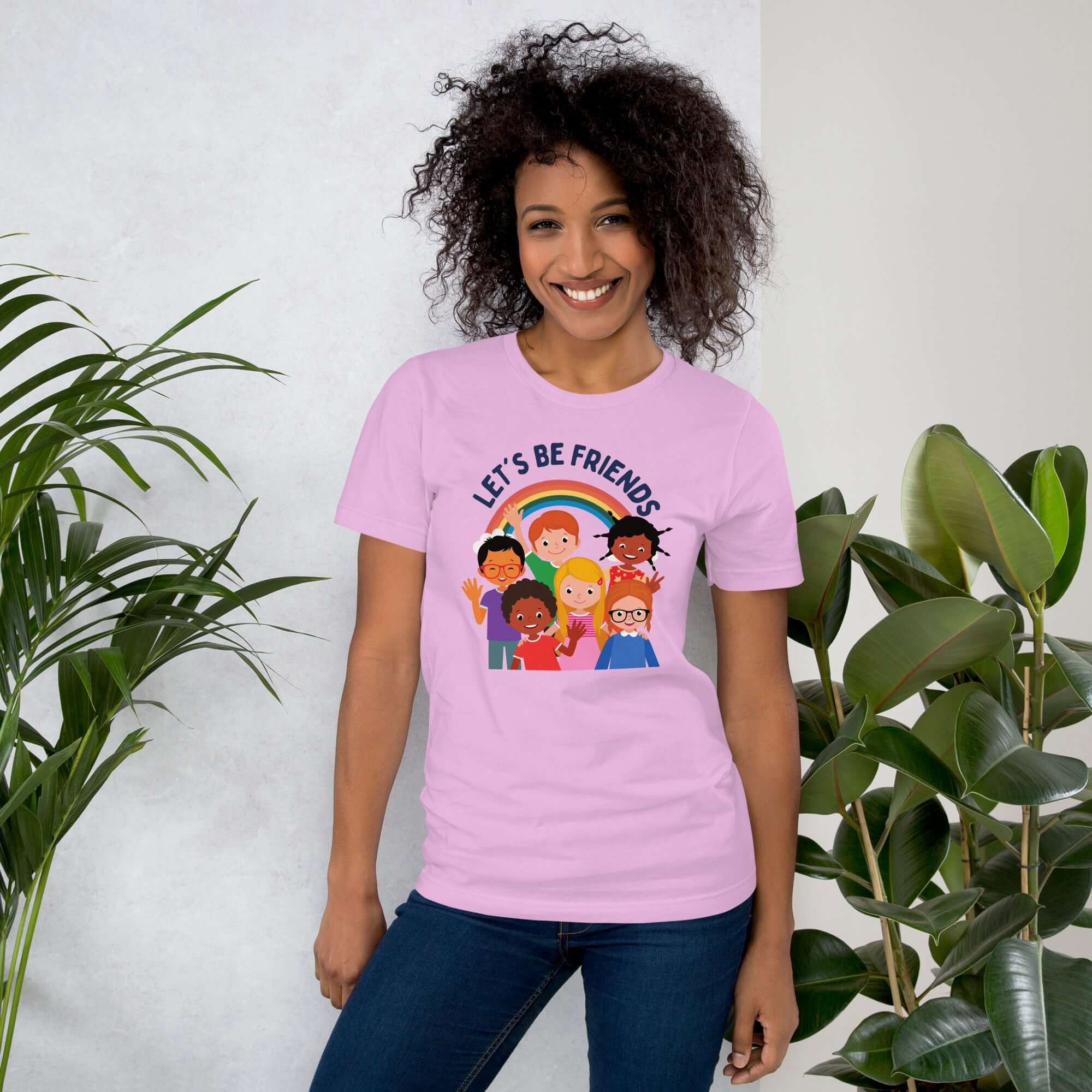 Smiling woman wearing a lilac Milk Sandwich t-shirt with a colorful "LET'S BE FRIENDS" graphic featuring six diverse cartoon kids and a rainbow, standing indoors near leafy green plants.