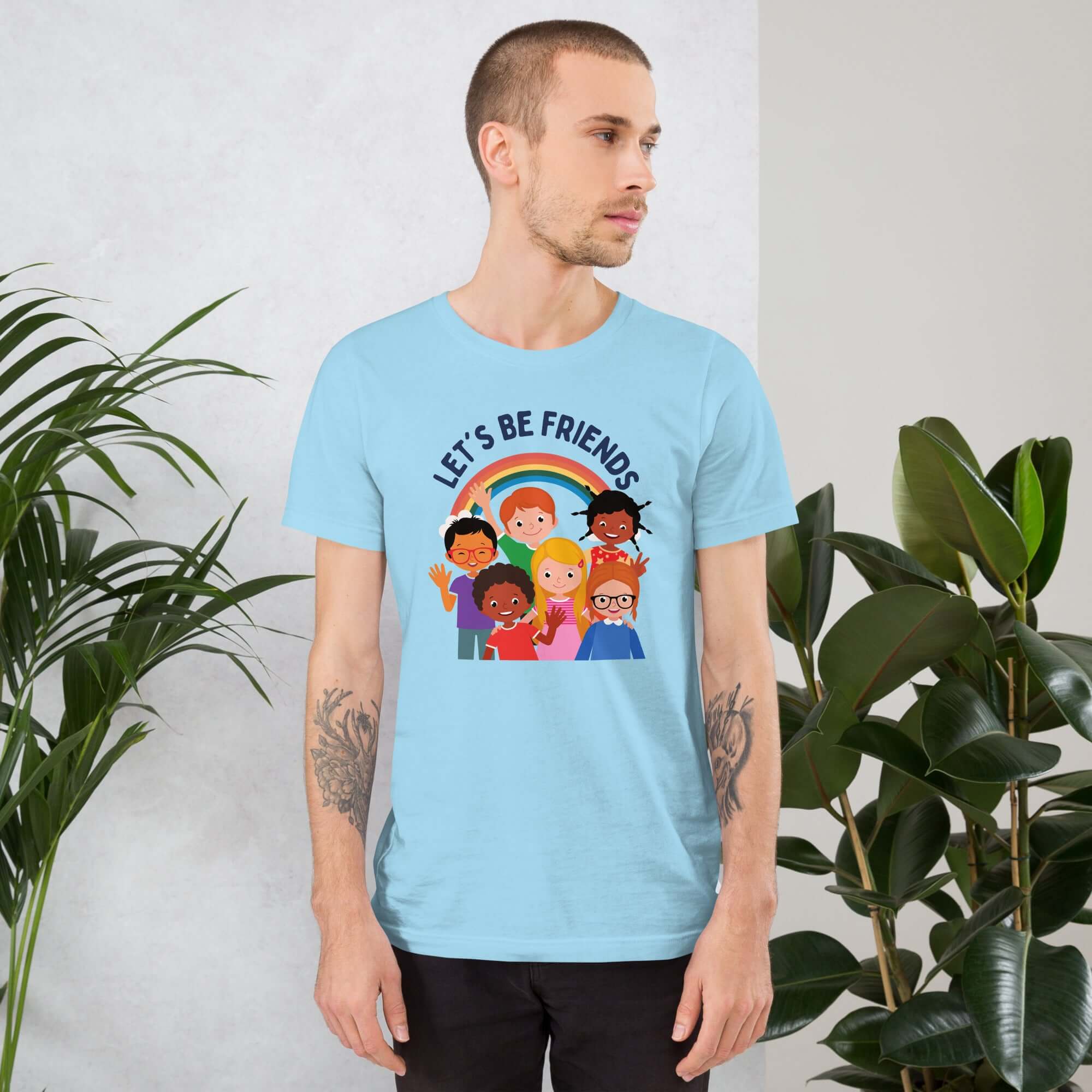 Man wearing a ocean blue Milk Sandwich t-shirt featuring a colorful illustration of six diverse cartoon kids smiling under a rainbow with the text "LET'S BE FRIENDS" standing indoors next to large green plants.