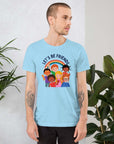 Man wearing a ocean blue Milk Sandwich t-shirt featuring a colorful illustration of six diverse cartoon kids smiling under a rainbow with the text "LET'S BE FRIENDS" standing indoors next to large green plants.